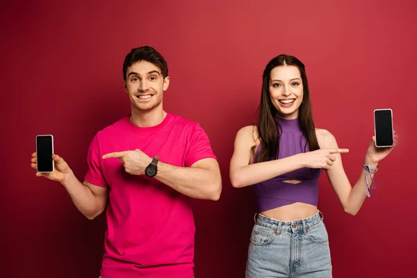 Beautiful smiling couple pointing at smartphones with blank screens on red — Stock Photo