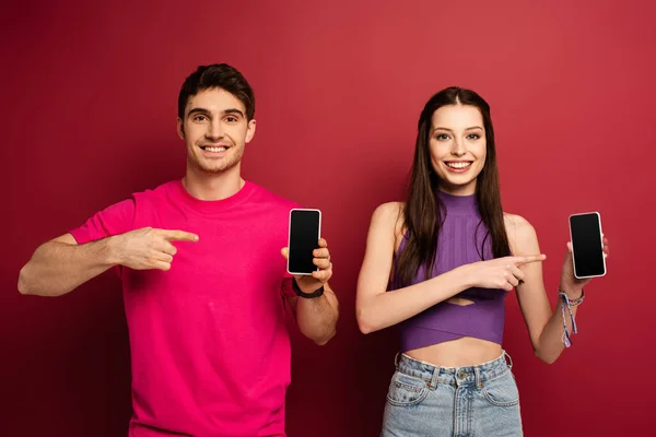 Couple souriant pointant vers les smartphones avec des écrans vides sur rouge — Photo de stock