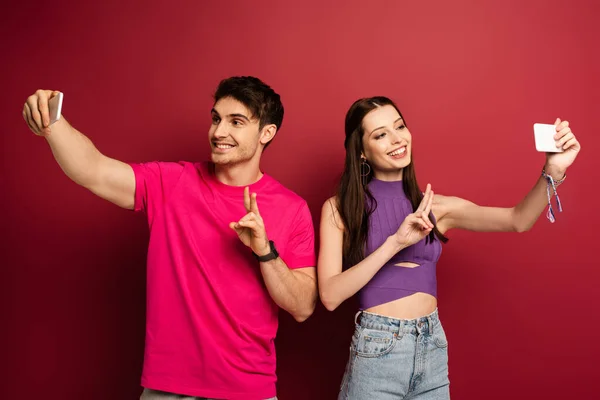 Smiling couple showing victory signs while taking selfie on smartphones on red — Stock Photo