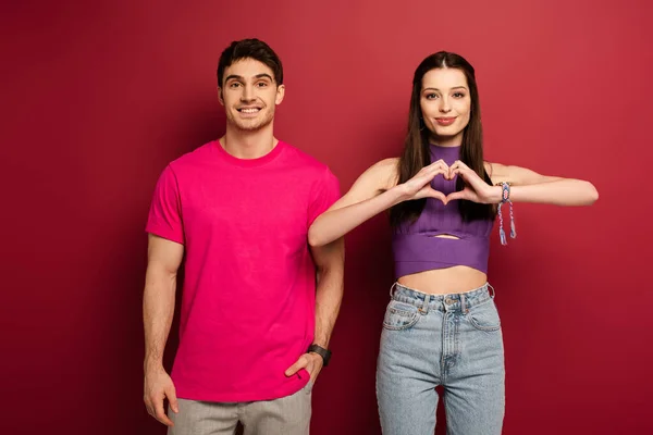 Positive man and beautiful girlfriend making heart sign with hands on red — Stock Photo