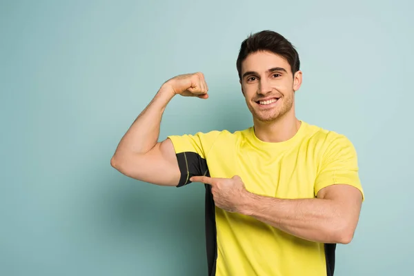 Smiling sportsman in yellow t-shirt pointing at muscles on blue — Stock Photo