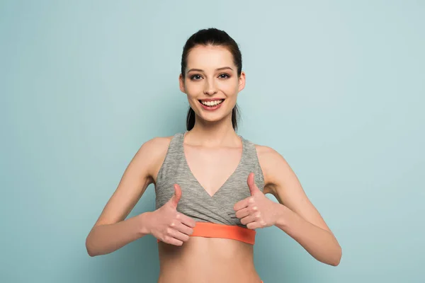 Beautiful happy athletic woman standing in sportswear and showing thumbs up on blue — Stock Photo