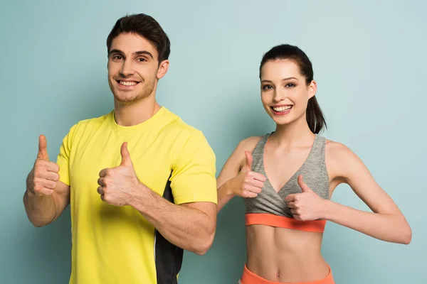 Feliz pareja atlética de pie en ropa deportiva mostrando pulgares hacia arriba en azul - foto de stock