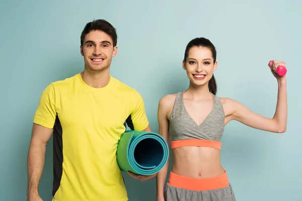 Cheerful athletic couple holding fitness mat and dumbbell on blue — Stock Photo