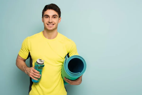 Desportista muscular alegre em t-shirt amarela segurando tapete de fitness e garrafa de esportes com água no azul — Fotografia de Stock