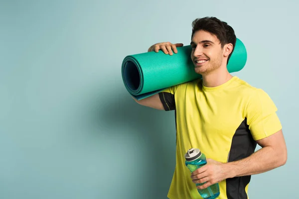 Sportivo muscoloso sorridente in t-shirt gialla con materassino fitness e bottiglia sportiva con acqua su blu — Foto stock