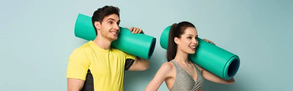 Panoramic shot of smiling athletic couple holding fitness mats on blue — Stock Photo