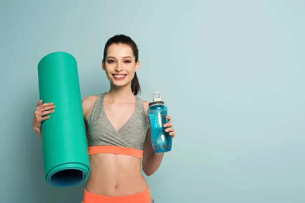 Улыбающаяся спортсменка держит спортивный коврик и спортивную бутылку с водой на голубом — стоковое фото