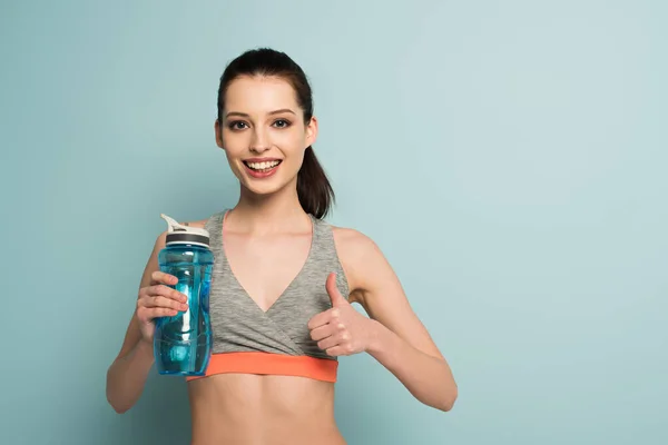 Glückliche sportliche Frau hält Sportflasche mit Wasser in der Hand und zeigt Daumen hoch auf blau — Stockfoto