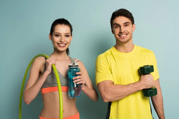 Feliz pareja atlética sosteniendo mancuerna, aro hula y botella de agua en azul - foto de stock