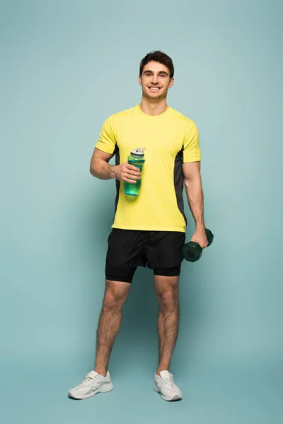 Sonriente deportista guapo en camiseta amarilla sosteniendo mancuerna y botella de deportes con agua en azul - foto de stock