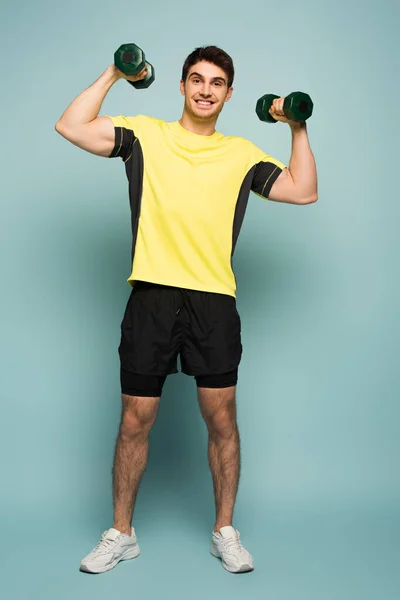 Cheerful muscular sportsman in yellow t-shirt training with dumbbells on blue — Stock Photo