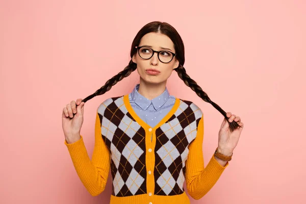 Thoughtful female nerd in eyeglasses holding braids on pink — Stock Photo