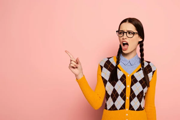 Shocked shouting female nerd in eyeglasses pointing on pink — Stock Photo