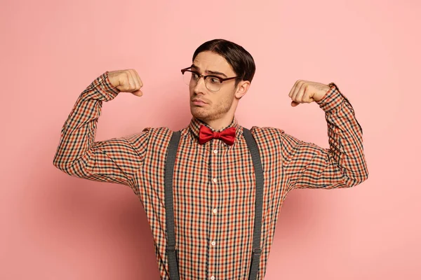 Thoughtful male nerd in eyeglasses showing muscles on pink — Stock Photo