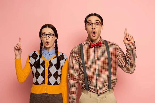 Couple of surprised nerds in eyeglasses having idea and pointing up on pink — Stock Photo