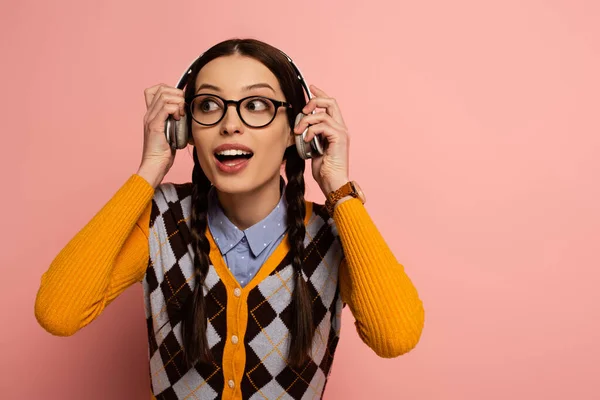 Nerd femenino sorprendido en gafas escuchando música con auriculares en rosa - foto de stock