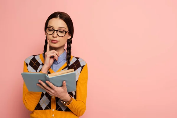 Pensativo nerd femenino en gafas lectura libro en rosa - foto de stock