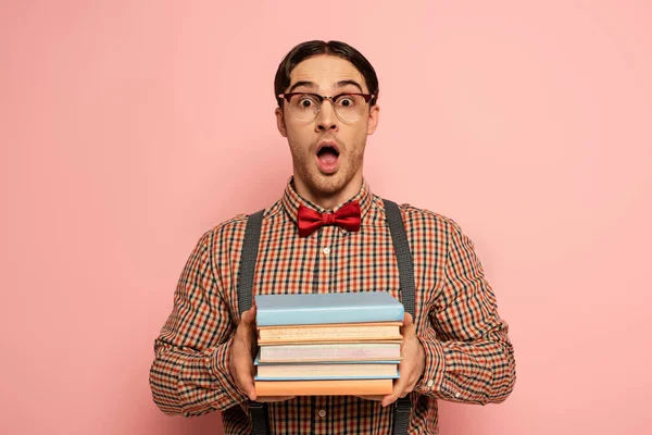 Nerd mâle choqué avec bouche ouverte dans des lunettes tenant des livres sur le rose — Photo de stock