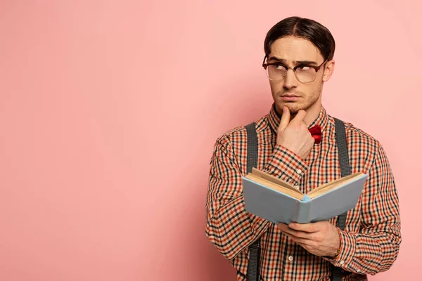 Thoughtful male nerd in eyeglasses reading book on pink — Stock Photo
