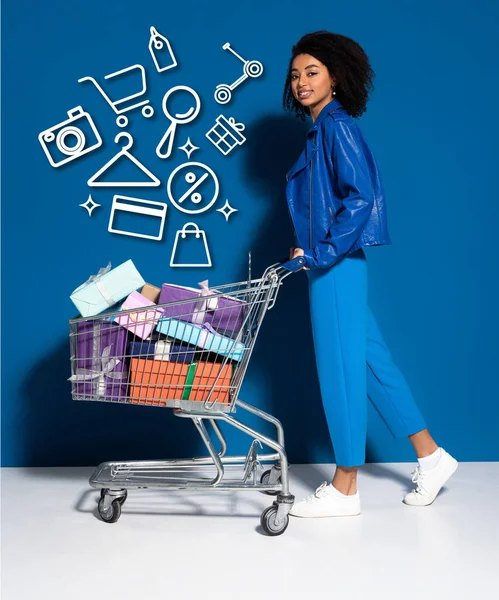 Side view of happy african american woman with shopping cart full of gifts on blue background — Stock Photo