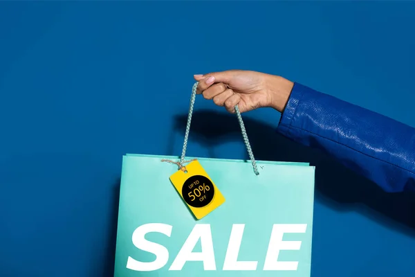 Cropped view of african american woman holding shopping bag with label on blue, 50 percent sale illustration — Stock Photo