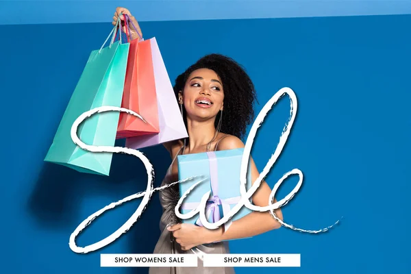 Happy beautiful african american woman in silver dress holding gift box and shopping bags while looking away on blue background, sale illustration — Stock Photo