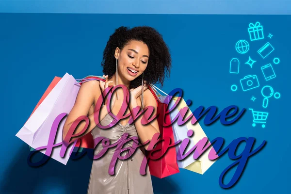 Elegante mujer afroamericana sonriente en vestido de plata sosteniendo bolsas de compras sobre fondo azul con ilustración de compras en línea - foto de stock