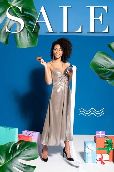 Щаслива красива афроамериканська жінка в срібній сукні тримає платіжний термінал з чековою та кредитною карткою біля подарунків та сумки для покупок на синьому фоні, продається ілюстрація — стокове фото