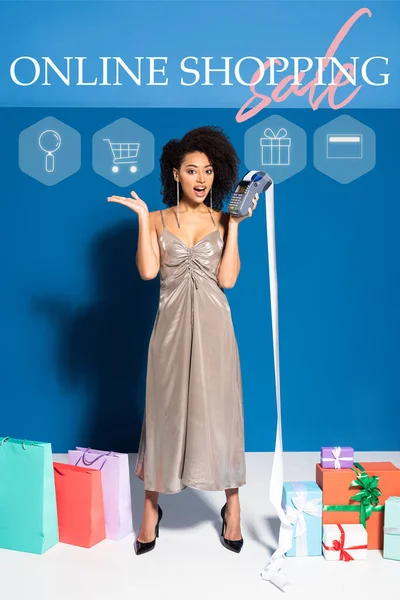Chocado bela mulher afro-americana em prata vestido segurando terminal com cheque perto de sacos de compras e presentes em fundo azul com ilustração de compras on-line — Fotografia de Stock