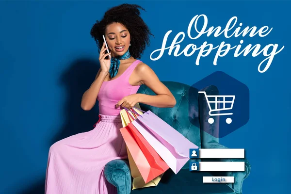 Sonriente mujer afroamericana sentada en un sillón de terciopelo con compras y hablando en un teléfono inteligente sobre fondo azul con ilustración de compras en línea - foto de stock