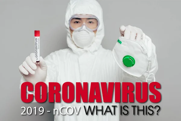 Asian epidemiologist in hazmat suit looking at camera while holding respirator mask and test tube with blood sample on grey background, coronavirus illustration — Stock Photo