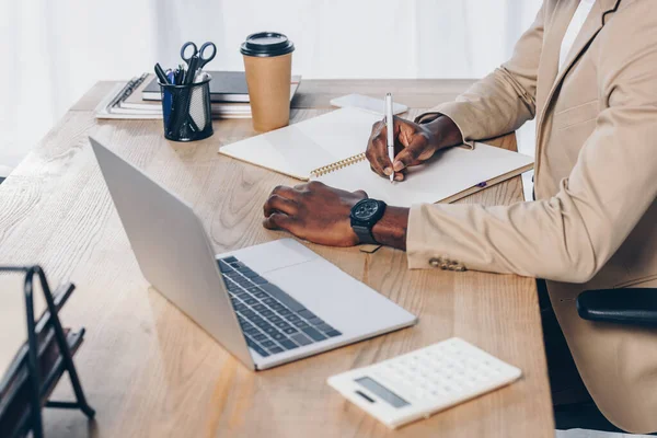 Ausgeschnittene Ansicht eines afrikanisch-amerikanischen Personalvermittlers, der in einem Notizbuch neben Laptop und Taschenrechner am Tisch im Büro schreibt — Stockfoto