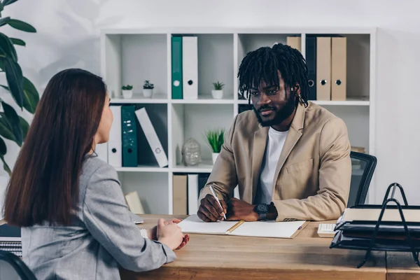 Reclutador afroamericano con bolígrafo y cuaderno realizando entrevista de trabajo con empleado en oficina - foto de stock