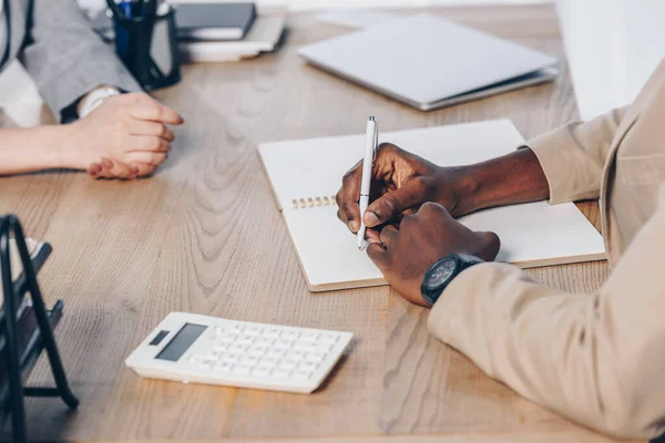 Vista cortada do recrutador realizando entrevista de emprego com empregado e escrevendo em caderno à mesa no escritório — Fotografia de Stock