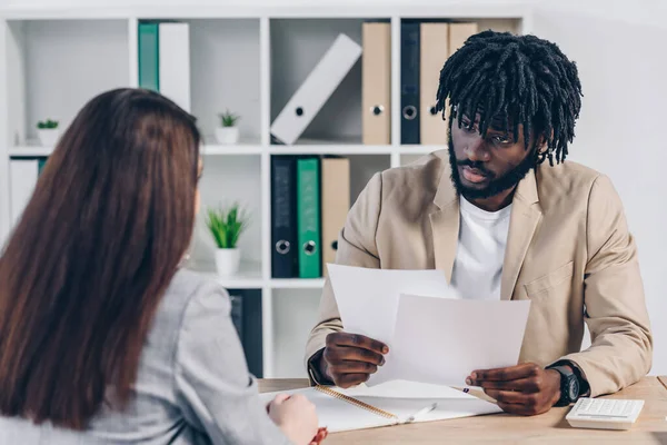 Enfoque selectivo del reclutador afroamericano sosteniendo papeles y mirando al empleado en la mesa en la oficina - foto de stock