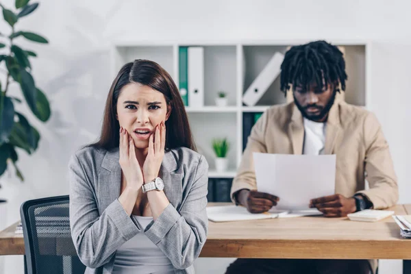 Scared employee looking at camera and african american recruiter reading documents at table in office — Stock Photo