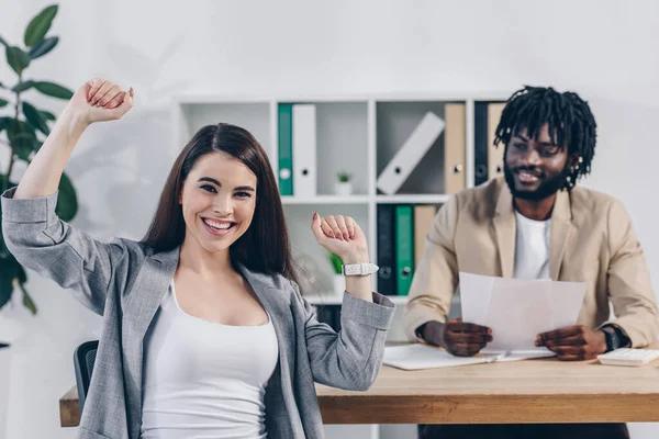 Excited employee looking at camera and african american recruiter with papers at table in office — Stock Photo