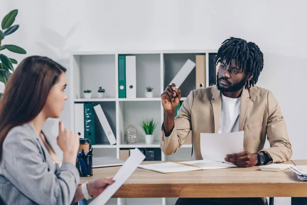 Selective focus of african american recruiter holding pen and looking at employee with papers at table in office — Stock Photo