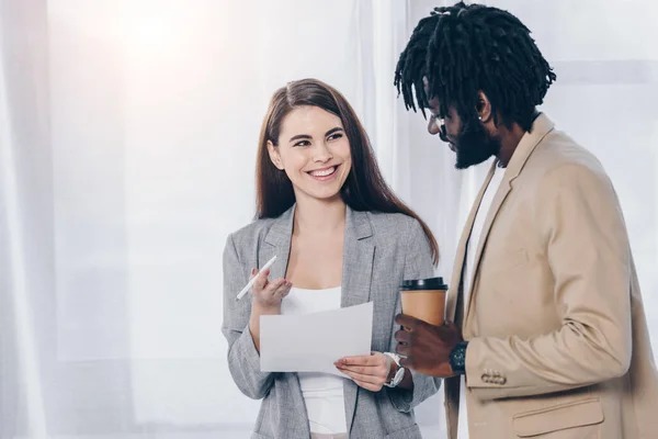 Recruiter with pen and paper looking at african american employee with disposable cup of coffee and smiling — Stock Photo