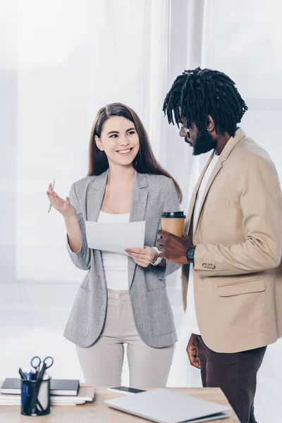 Recrutador com caneta e papel olhando para empregado americano africano com xícara descartável de café e sorrindo perto da mesa — Fotografia de Stock