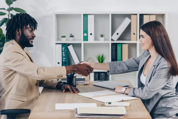 Recrutador afro-americano e empregado apertando as mãos, olhando um para o outro e sorrindo no escritório — Fotografia de Stock