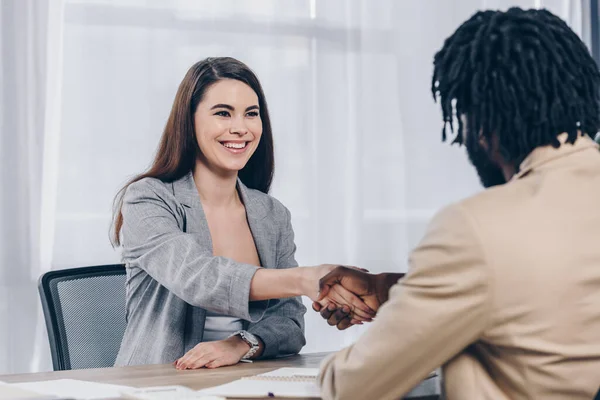 Selective focus of employee smiling and shaking hands with african american recruiter at job interview in office — Stock Photo