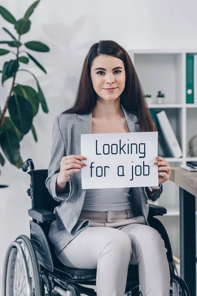 Dipendente disabili sorridente, guardando la fotocamera e tenendo cartello con la ricerca di un lavoro lettering sulla sedia a rotelle in ufficio — Foto stock