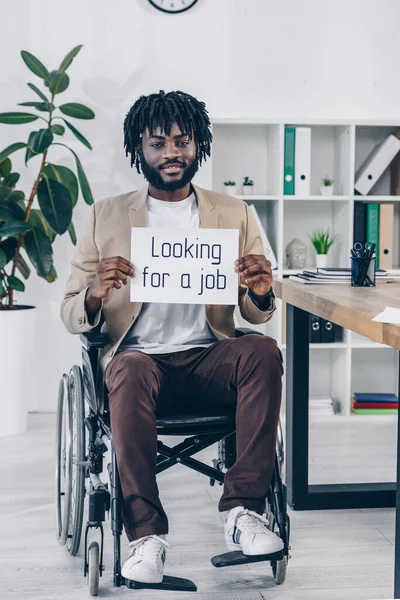 Empregado afro-americano deficiente segurando cartaz com a procura de emprego lettering em cadeira de rodas no escritório — Fotografia de Stock