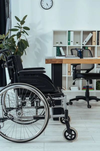 Black wheelchair near table and plant in office — Stock Photo