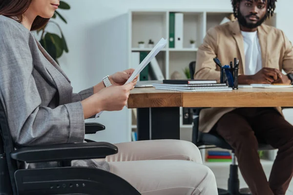 Empleado discapacitado en silla de ruedas con papeles y reclutador afroamericano en la mesa en la oficina - foto de stock