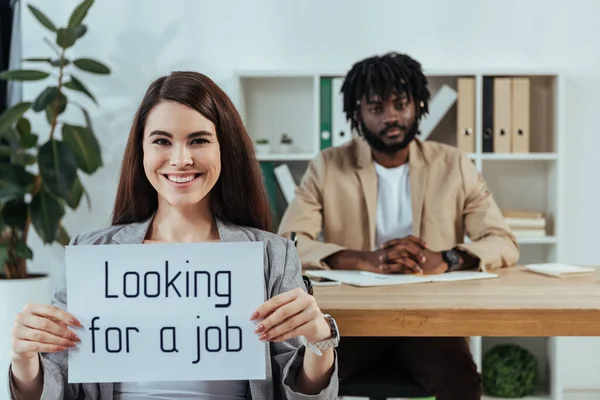 Empleado mostrando pancarta con la búsqueda de un puesto de trabajo y reclutador afroamericano en la oficina - foto de stock