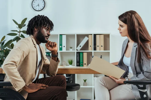 Empleado afroamericano discapacitado y reclutador con carpeta mirándose en la entrevista de trabajo en la oficina - foto de stock