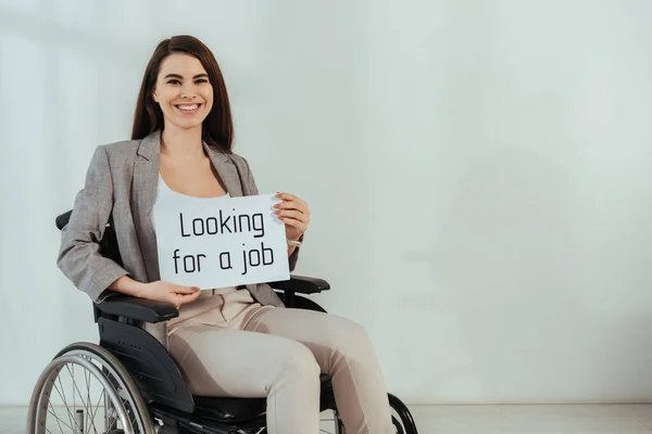 Жінка з обмеженими можливостями посміхається і тримає плакат з пошуком роботи на інвалідному візку на білому тлі — стокове фото